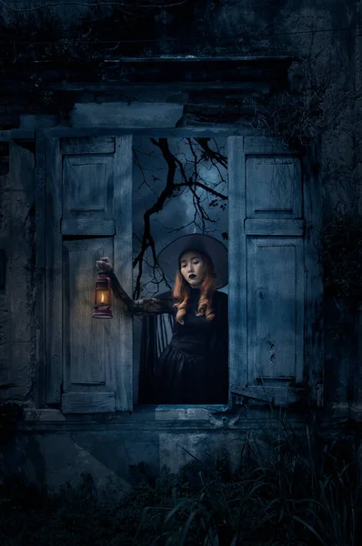 死んだ木と不気味な曇りの空 ハロウィーンの謎の概念の上に壁と古い破損した木の窓に立っている古代のランプを保持するハロウィーンの魔女 — ストック写真