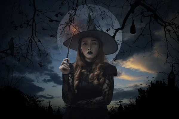 Halloween witch holding magic wand standing over cross, church, crow, bat, birds, dead tree, full moon and sunset sky, Halloween mystery concept
