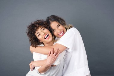 Beyaz tişört giyen, gülümseyen ve stüdyoda gri arka planda sarılan iki güzel beyaz kadın. 