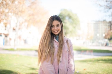 Güven ve iş kavramı. Pembe ceketli, açık havada poz veren, kendinden emin bir şekilde gülümseyen çekici sarışın kadın portresi.