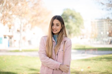 Güven ve iş kavramı. Pembe ceketli, açık havada poz veren, kendinden emin bir şekilde gülümseyen çekici sarışın kadın portresi.