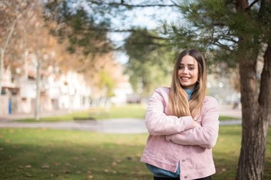 Açık havada kolları bağlı pembe ceket giyen neşeli, yeşil gözlü genç bir kadının portresi. Güzel beyaz kız kollarını kavuşturup kameraya bakıp gülümsüyor.