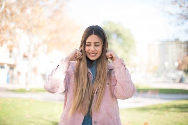 Pembe ceketli güzel sarışın kadın parkta kulaklarını kapatıyor.  