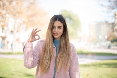İyi iş çıkardın. Mutlu gülümseyen mavi gözlü, pembe ceket giyen başarılı genç bir kadının portresi. Açık havada güzel bir jest yapıyor. Olumlu insani duygu yüz ifadesi vücut dili. Komik kız.