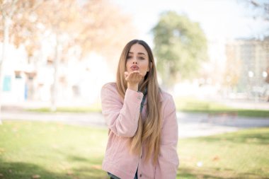 Pembe ceketli, modern saçlı, sevimli sarışın bir kadının portresi. Açık havada kameraya bakarak dudak bükerek öpücük yolluyor. Duygular konsepti