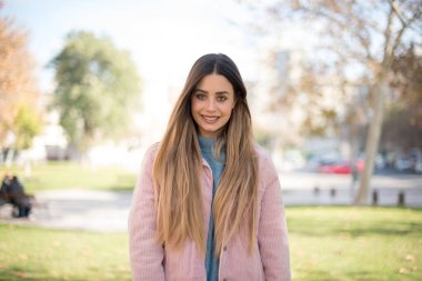 Pembe ceket giyen neşeli sarışın kadın ellerini aşağıda tutuyor ve gülümsüyor ve kendinden emin, gündelik kıyafetler giyiyor ve güçlü bir görünüm sergiliyor. Mutlu genç kadın.