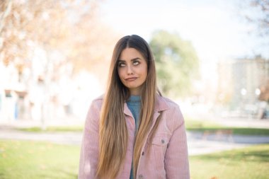 Genç, güzel, sarışın, pembe ceketli bir kadın parkta komik bir ifade takınıyor.  