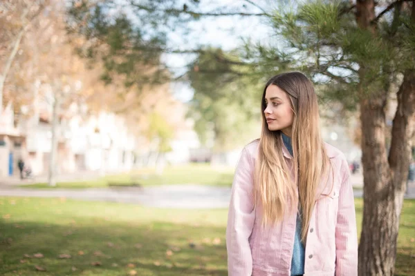 Pembe ceket giyen neşeli sarışın kadın ellerini aşağıda tutuyor ve gülümsüyor ve kendinden emin, gündelik kıyafetler giyiyor ve güçlü bir görünüm sergiliyor. Mutlu genç kadın.