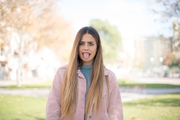 年轻美丽的金发女子穿着粉色夹克 在公园里露出舌头 — 图库照片