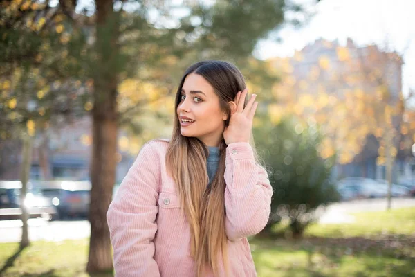若い美しいブロンドの女性は公園で聞いているピンクのジャケットを着ています — ストック写真