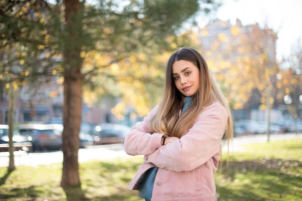 Açık havada kollarını kavuşturmuş pembe ceket giyen neşeli sarışın bir kadın resmi. Kameraya bakıp gülümsüyor. Kendine güvenen kız.
