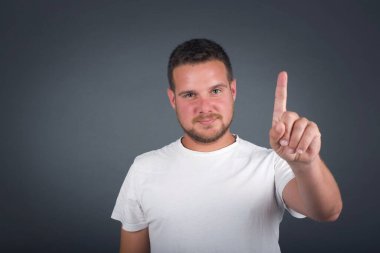 Kafkasyalı genç adam gri duvarın önünde duruyor. Bir numaralı parmaklarıyla işaret ediyor ve gülümsüyor. Kendinden emin ve mutlu..