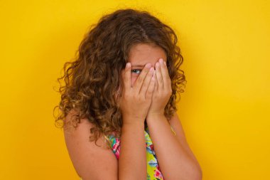 Endişe - yazlık elbise giyen, sarı arka planda yüzünü elleriyle örten ve parmaklarının arasında duran bir gözüyle dışarı bakan güzel bir kızın kavramsal görüntüsü. Bir şeyden ya da birinden korktun mu?.