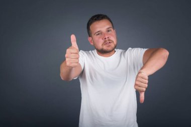 Oldukça düşünceli genç bir adam iyiye kötüye işaret yapıyor. Keyifsiz ve etkilenmemiş sıradan kıyafetler giyip gri duvarın önünde dikiliyor..