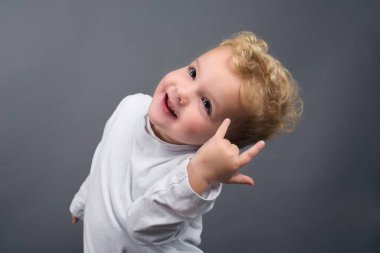 Sarı kıvırcık saçlı şirin çocuk stüdyoda gri arka planda şeytanın boynuzlarını gösteriyor.