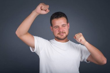 Çekici beyaz bir adam zaferi kutluyor yumruklarıyla havaya yumruk atıyor ve gri bir stüdyo arka planında ışıltılı bir gülümseme ile fotokopi çekiyor.