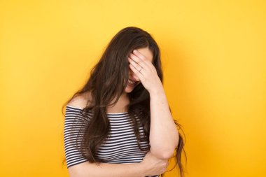 Havalı saç kesimi olan, günlük kıyafetler giyen, gülümseyen, sarı arka planda duran, aptalca durumlara şaşırmış, el hareketi yapan güzel bir kadının kapalı bir portresi..