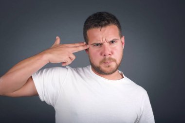 Mutsuz bir adam silah atışı taklidi yapıyor intihar jesti yapıyor iki parmağını şakakların üstünde tutuyor günlük kıyafetleri giymiş şaşkın ifade içeride duruyor..