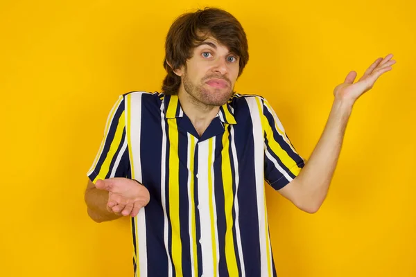 handsome caucasian young male pointing aside with both hands showing something strange and saying: I don\'t know what is this. Standing against gray background. Advertisement concept.