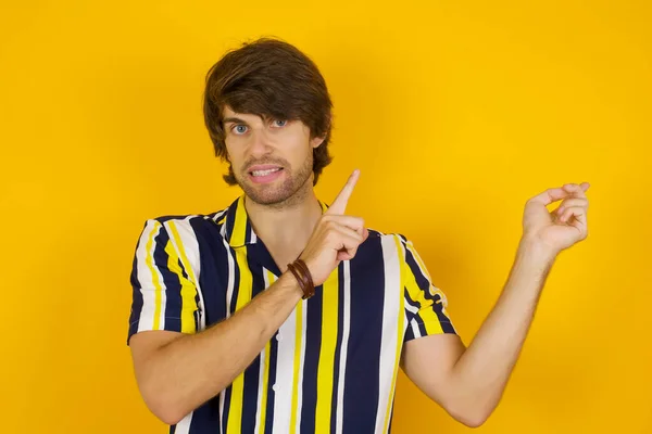 Pretty young man with positive expression, dressed casually, indicates with fore finger at blank copy space for your promotional text or advertisement. Adorable man poses indoor alone