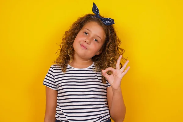 Glad attractive woman shows ok sign with hand as expresses approval, has cheerful expression, being optimistic. Standing against white wall.