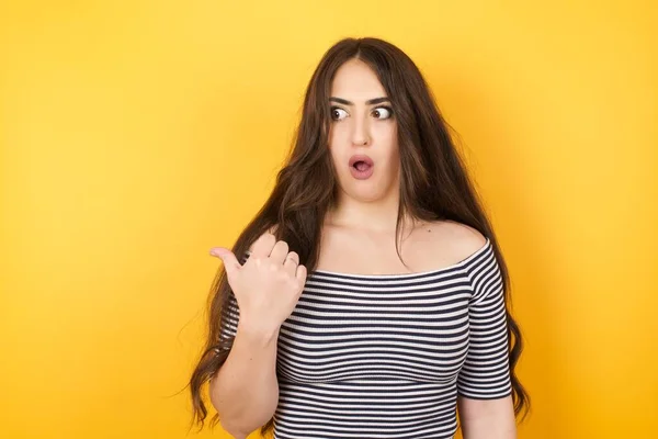 Horizontal shot of pretty young joyful shocked  woman in casual clothes  points with thumb away, indicates something on blank wall isolated over white background. Check this out. Ethnicity concept.