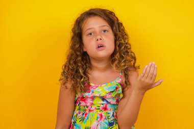 What the hell are you talking about, nonsense. Studio shot of frustrated little girl wearing summer dress on yellow background with blonde hair gesturing with raised palm, frowning, being displeased and confused with dumb question