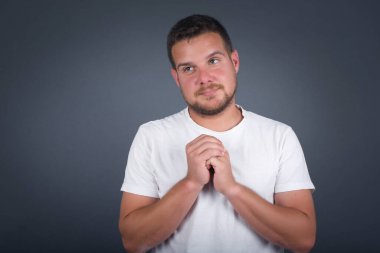 Tatlı yüzlü, rüya gibi, genç bir erkek, ellerini yüzünün yanında çapraz tutuyor, hoş bir şey için heyecanlı, gri arka plana karşı duruyor..