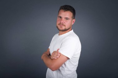 Beyaz tişört giymiş, kolları çapraz gri duvarda duran neşeli beyaz bir adam resmi. Kameraya bakıp gülümsüyor. Kendine güvenen başarılı bir adam..