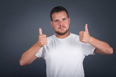 Gri arka planda izole edilmiş, olumlu içerik ve mutluluk duygularıyla baş parmaklarını gösteren iyimser genç adama yakın çekim. Ortak alan, kalite ve tavsiye ile tatmin kavramı.