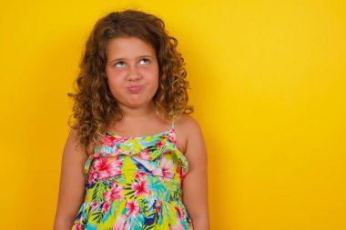 Kasvetli, sarı arka planda yaz elbisesi giyen küçük bir kız dudaklarıyla yukarıya bakıyor, önemli bir şey hakkında düşünerek üzülüyor, ellerini aşağıda tutuyor..