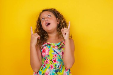 Sarı arka planda, parlak makyajlı, yaz elbisesi giymiş, her iki işaret parmağını yukarı kaldırıp boş fotokopi duvarında şok edici bir şey gösteren başarılı, dostane görünümlü küçük bir kızın yatay çekimi..