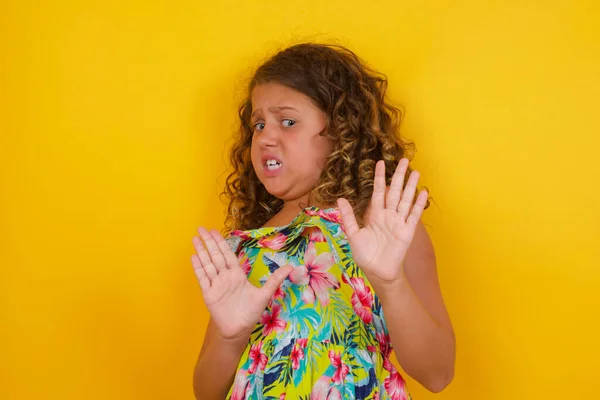 Dissatisfied little girl wearing summer dress on yellow background frowns face, has disgusting expression, shows tongue, expresses non compliance, irritated with somebody, rejects do something. People and negative facial expressions