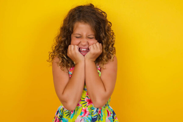 Portrait of young little girl wearing summer dress on yellow background being overwhelmed with emotions, expressing excitement and happiness with closed eyes and hands near face while smiling