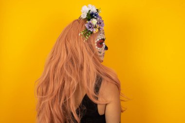 Cadılar Bayramı makyajlı, uzun düz dalgalı ve parlak saçlı sarı duvarda duran bir kadının arka görünüşü. Stüdyo Çekimi.