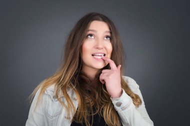 Düşünceli bir ifadeyle hayalperest beyaz kadın, kameraya bakıyor, elini yüzüne yakın tutuyor, parmağını ısırıyor hoş bir şey düşünüyor, gri duvara karşı poz veriyor..
