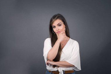 Düşünceli gülümseyen kızın portresi elini çenesinin altında tutuyor, doğrudan kameraya bakıyor, ilgiyle karışık bir şey dinliyor, sıradan giyiniyor, gri duvara karşı poz veriyor. Gençlik kavramı.