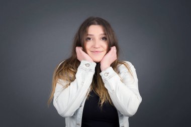 Hayalperest genç Avrupalı kadın ellerini çenesinin altına bastırıyor, mutlu bir ifadeyle bakıyor, dişlek gülüşü var, gri arka planda soyutlanmış.