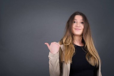 Omg konsepti. Şaşırmış beyaz kadın, şaşırmış bir ifadeyle, gözlerini ve ağzını iyice açıyor, baş parmağıyla işaret ediyor, gri arka planda garip bir şey gösteriyor. Reklam kavramı.