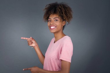 Pozitif çekici genç Afrikalı Amerikalı kadın gri stüdyoda dururken neşeli ve şaşırmış bir ifadeye işaret ediyor.