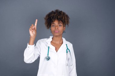 Genç Afrikalı Amerikalı doktor üniformalı kadın gri duvarın önünde duruyor ve parmaklarını bir numara ile gösterirken kendinden emin ve mutlu gülümsüyor..