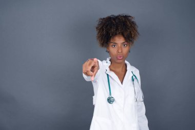 Kapalı alanda çekilen Afro-Amerikan kadın doktor, size mutlulukla işaret ediyor, rekabeti seçiyor, olumlu ifade ediyor, tercihini yapıyor, gri duvarda izole edilmiş bir şekilde..