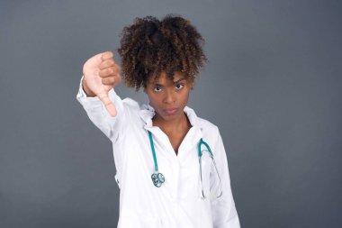 Hoşnutsuz Afrikalı Amerikalı doktor kadın onaylamadığını, başparmağını aşağıda tuttuğunu, hoşlanmadığını ifade ettiğini, hoşnutsuzca kaşlarını çattığını, gri arka planda soyutlandığını gösteriyor. Vücut dili kavramı.