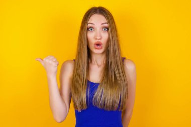 Oldukça genç ve neşeli bir kadının yatay görüntüsü başparmağıyla işaret ediyor. Bu da boş duvarda sarı arkaplanda izole edilmiş bir şey olduğunu gösteriyor. Şuna bir bak. Etnik konsept.