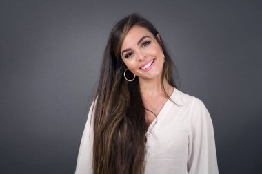 Geniş tebessümlü güzel esmer kadın, beyaz dişlerini gösteriyor, günlük tişört giyiyor, gri stüdyo duvarının başında dikiliyor izin gününün keyfini çıkarıyor. Kadın içeride duruyor..