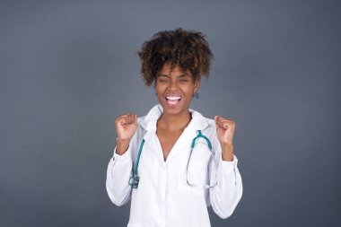  Afro-Amerikalı kadın doktor üniforma giyiyor mutlu ve kazanan jest için heyecanlanıyor. Başarılı ve zaferi kutlayan, muzaffer, gri arka plan.