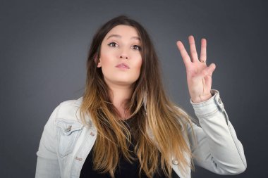 Genç, orta yaşlı, beyaz bir kadın gri duvarın önünde duruyor. Üç numaralı parmaklarıyla işaret ediyor. Kendinden emin ve mutlu bir şekilde gülümsüyor..