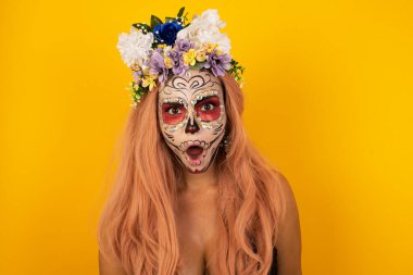 Etkileyici yüz ifadeleri. Şok olmuş, cadılar bayramı makyajlı güzel bir kadın çenesini kapatıyor, boş sarı duvarda gördüklerinden sersemlemiş hissediyor.