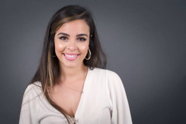 Mutlu ve komik yüzlü, gülümseyen ve dil gösteren güzel beyaz bir kadın. Günlük giysiler giyip gri stüdyonun arka planında dikiliyorlar..