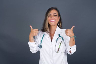 Gri arka planda izole edilmiş, olumlu içerik ve mutluluk duygularıyla baş parmaklarını gösteren genç iyimser doktorun yakın çekimi. Kalite ve tavsiye ile tatmin kavramı.